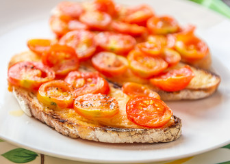 Grilled sliced fresh cherry tomatoes on buttered toasted sough dough bread on a white plate. Below is a tray with a hint of its leafy pattern. Light with natural light.