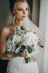 Fototapeta premium Stylish bride holding modern wedding bouquet and posing in soft light near window in hotel room. Gorgeous sensual bride portrait. Morning preparation before wedding ceremony