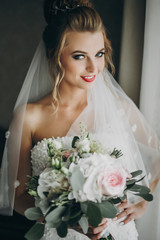 Naklejka premium Stylish bride holding modern wedding bouquet and posing in soft light near window in hotel room. Gorgeous sensual bride portrait. Morning preparation before wedding ceremony