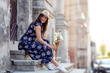 Beautiful woman with hat and flowers on hand sitting at the stairs in old town