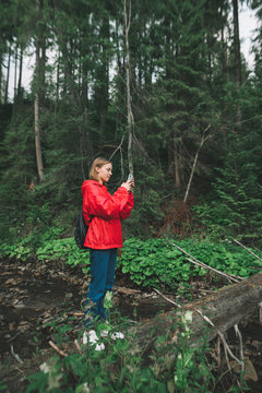 Attractive girl in a red raincoat stands in the forest on a background of a mountain stream, makes a photo on a smartphone. Girl traveler uses a smartphone on a hike, stands on a log over the river
