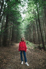 Obraz premium Young woman in a red jacket stands on a path in the forest, looks into the camera with a serious face. Portrait of hipster girl hiking through forest. Hiking mountains concept.