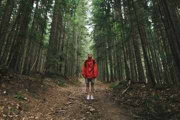 Obraz premium Hiker man in a red raincoat stands on a trail in the mountains,looks in camera, guy on a mountain hike. Full length photo of tourist in red jacket standing in background of big old coniferous trees