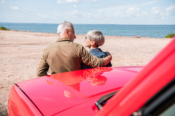 Fototapeta na wymiar back view of senior couple standing near red car at beach and embracing in sunny day
