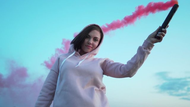 Young pretty woman with pink smoke bomb or grenade on sunset sky background.