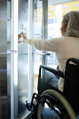 Senior female patient sitting in wheelchair and pushing the button of elevator she moving down