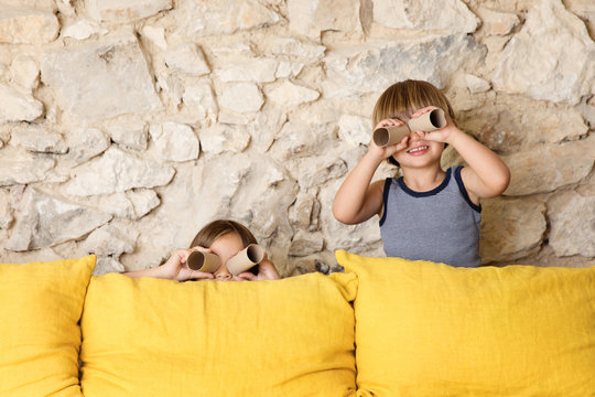 Two kids behind a yellow sofa looking through toilet paper rolls