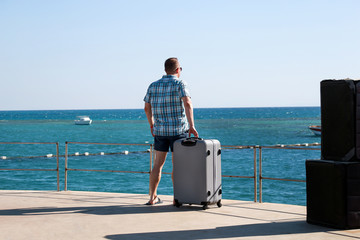 Fototapeta premium Tourist and traveler is stand on beach of hotel resort with traveled suitcase, enjoys view sea environment, nature landscape, panorama, horizon and rest in sun. Handsome man on vacation in summertime.