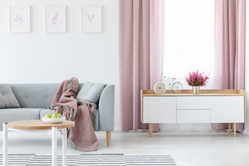 Blanket on grey couch in elegant living room interior with white wooden cabinet and coffee table, striped carpet and lilac curtains, real photo