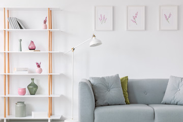 Prints in frames above grey stylish couch with pillows, white lamp and shelf with vases and books next to it, real photo