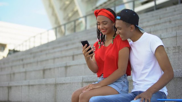 Couple of mixed-race teenagers sitting outdoor scrolling web pages on smartphone