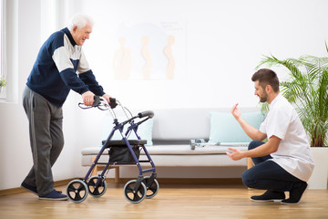 Elderly grandfather with walker trying to walk again and helpful male nurse supporting him - obrazy, fototapety, plakaty