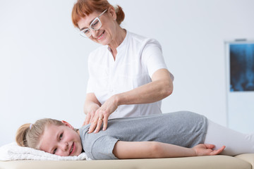 Positive senior physiotherapist massaging a cute blond girl with spine deformity during session in...