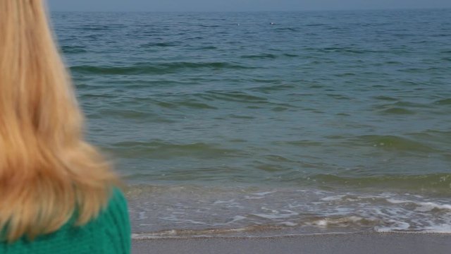 Closeup back view of blond beautiful woman sitting alone at beach isolated at blue sea water background. Focus at water. Real time full hd video footage.