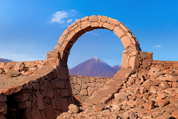 Desert landscape of volcano seen through arch in Pukara de Quitorand near San Pedro de Atacama, Chile, against a blue sky.