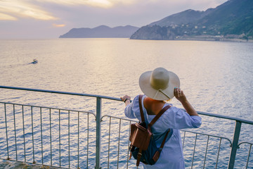 Woman in casual dress and hat with backpack looking to sea. Travel Cinque Terre in Italy in summer sunset