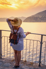 Woman in casual dress and hat with backpack looking to sea. Travel Cinque Terre in Italy in summer sunset