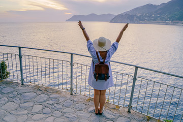 Woman in casual dress and hat with backpack looking to sea. Travel Cinque Terre in Italy in summer sunset