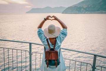 Woman in casual dress and hat with backpack looking to sea and making heart of hands. Travel Cinque Terre in Italy in summer sunset