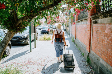 Woman in hat with suitcase and backpack is walking on city street in sunny summer day. Italy, Europe