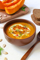 Roasted pumpkin and carrot soup with cream, black pepper and pumpkin seeds ,cutting board and fresh pumpkin slices,black bread on white concrete background.