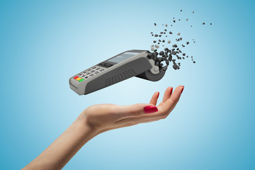 Closeup of woman's hand levitating point-of-sale terminal which has started to break into small pieces on light blue background.