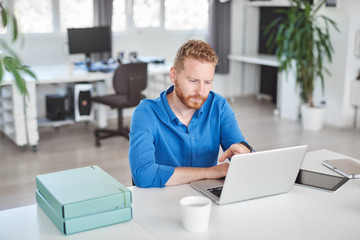 Young serious handsome Caucasian male employee sitting in office and using laptop. Start up...