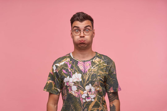 Indoor shot of cute guy in flowered t-shirt with wide eyes open standing over pink background, trying to hold breath, making funny grimace
