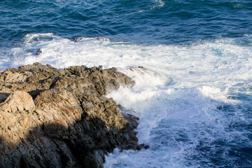 Powerful waves crashing into rocks in a mediterranean sea, blue water with foam.