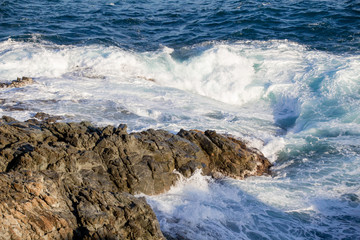 Powerful waves crashing into rocks in a mediterranean sea, blue water with foam.