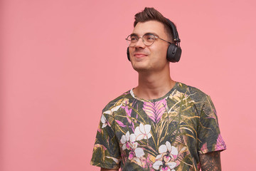 Cute tattooed guy wearing glasses and headphones, standing over pink background, listening to music with amused smile, looking happy and pleased