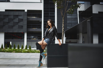 Beautiful and happy young model woman in the black leather jacket, in jeans shorts and stockings, smiling and posing at the modern urban background