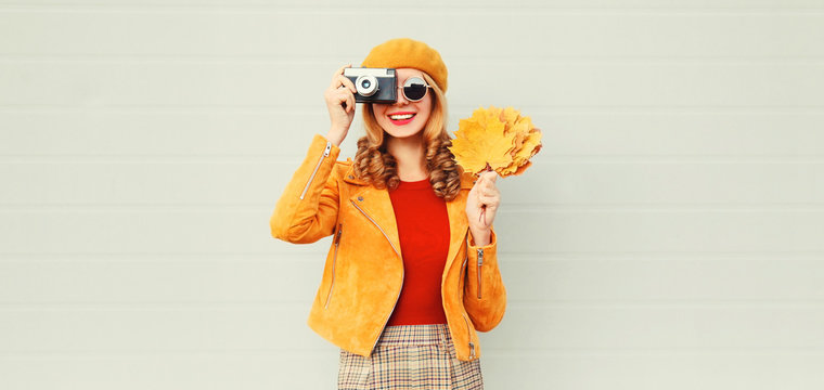 happy smiling woman with autumn yellow maple leaves, retro camera taking picture in french beret over gray wall background