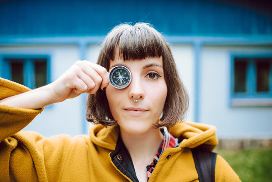 Cheerful young woman crossing eyes and holding retro compass near face while standing on blurred background of countryside house