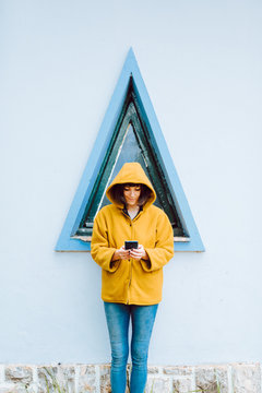Young woman in yellow warm coat smiling and looking down while standing against triangle window and gray wall of building