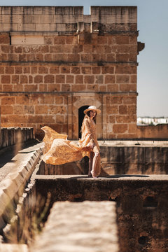Side view of trendy woman in hat and flying dress standing on deserted building by brick wall