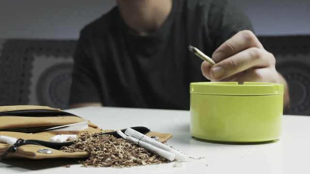 Pan shot of man's hands detail while smoking a handmade cigarette , tobacco holder and tabacco accessories on the table foreground