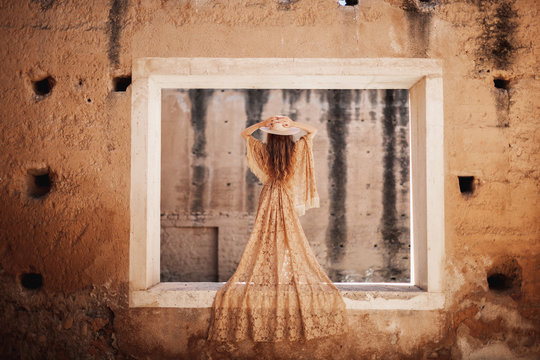Back view of gorgeous woman in beige lace dress standing on wall of deserted building and holding hat