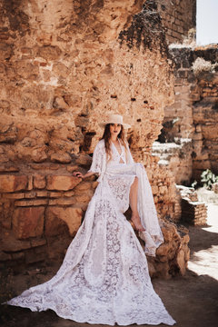 Beautiful woman in long white dress and hat sitting by wall of old building and looking at camera