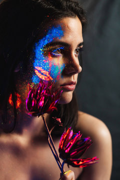 Portrait of beautiful young woman covered with luminous paint on face holding a flower and looking away.