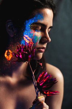 Portrait of beautiful young woman with closed eyes covered with luminous paint on face holding a flower
