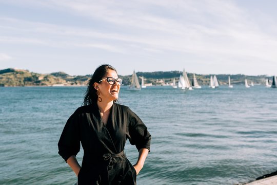 Side view of young attractive woman in black outfit standing by sea and leaning on pole in Lisbon