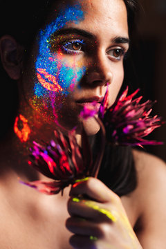 Portrait of beautiful young woman covered with luminous paint on face holding a flower and looking away.