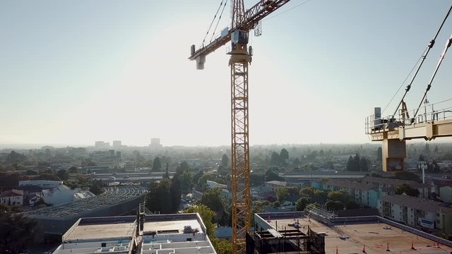 Two tower cranes at sustainable, urban construction site with sun and highway behind, Drone shot pulling out.