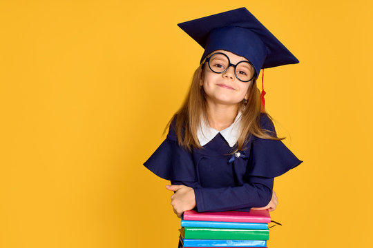 Cheerful schoolgirl in graduation outfit smiling while leaning on pile of colourful books over yellow background