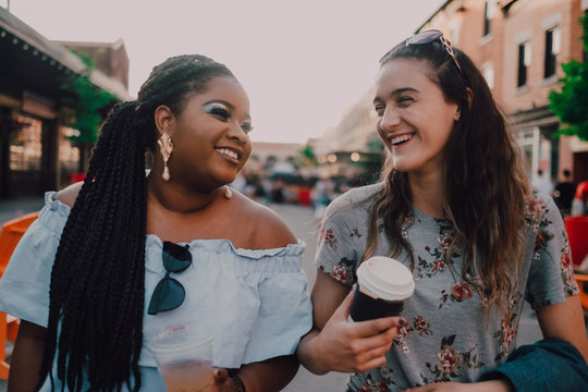 Cheerful trendy multiracial young casual women talking and drinking coffee while walking on street on sunset