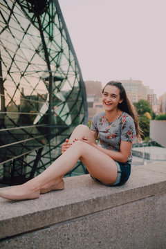 Calm content casual young woman in shorts and t-shirt enjoying sunshine while sitting on concrete parapet looking at camera