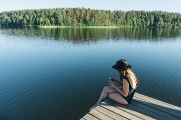 Side view of young woman in black swimsuit and hat sitting on wooden pier on the mobile phone on a lake on clear blue sky and forest background