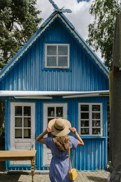 Back view of woman in straw hat and sundress standing in front of colourful blue summer house in daylight