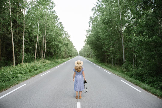 Back view of adult woman in straw hat and dress with camera standing in middle of drive way among tall green trees